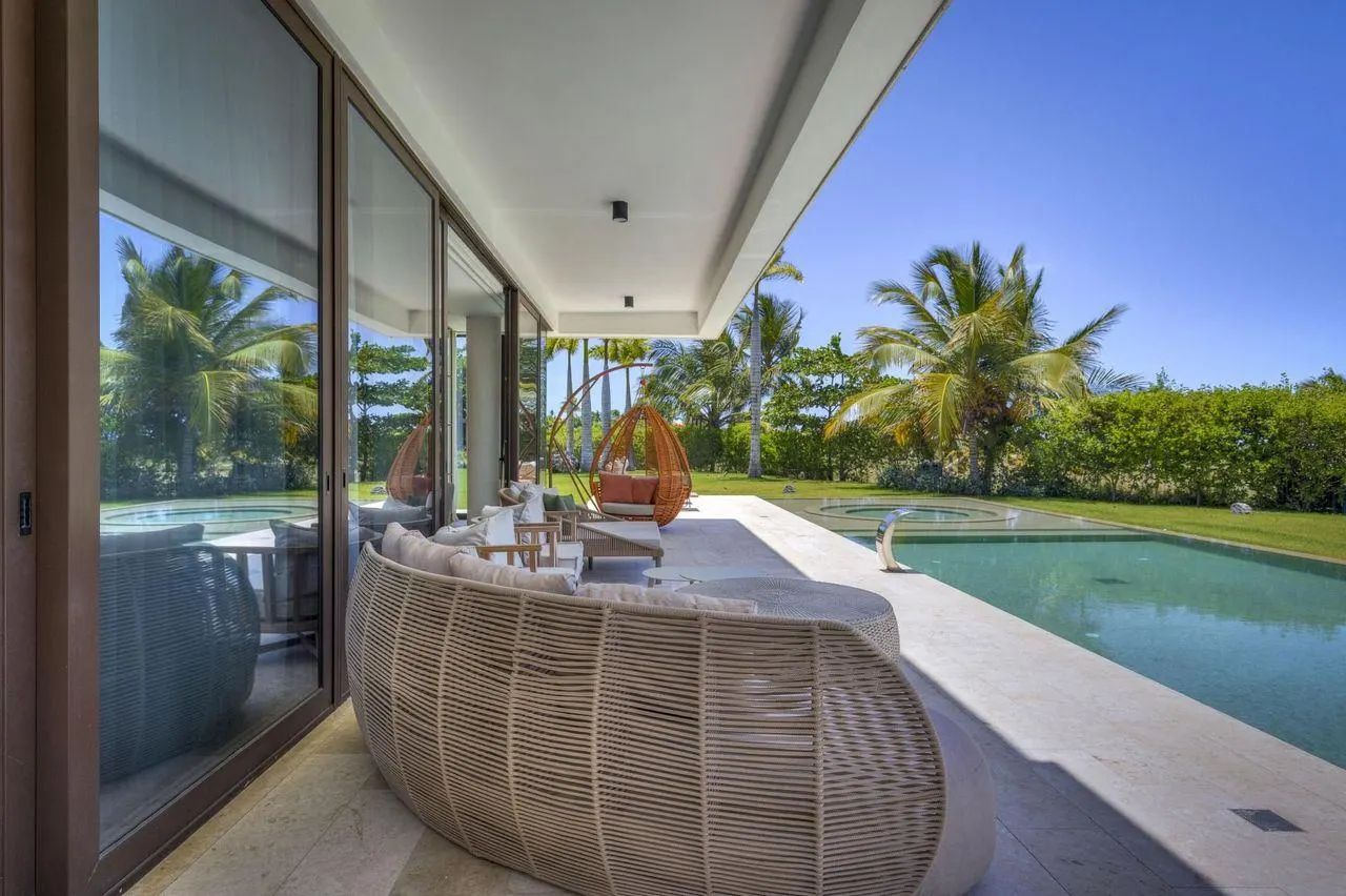 A covered seat by the glass, the pool and the garden beyond